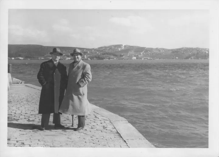 Kış ayında deniz kenarında şık paltoları ve şapkalarıyla poz veren iki erkek. Arka planda tepeler ve sahil şeridi yer alıyor.