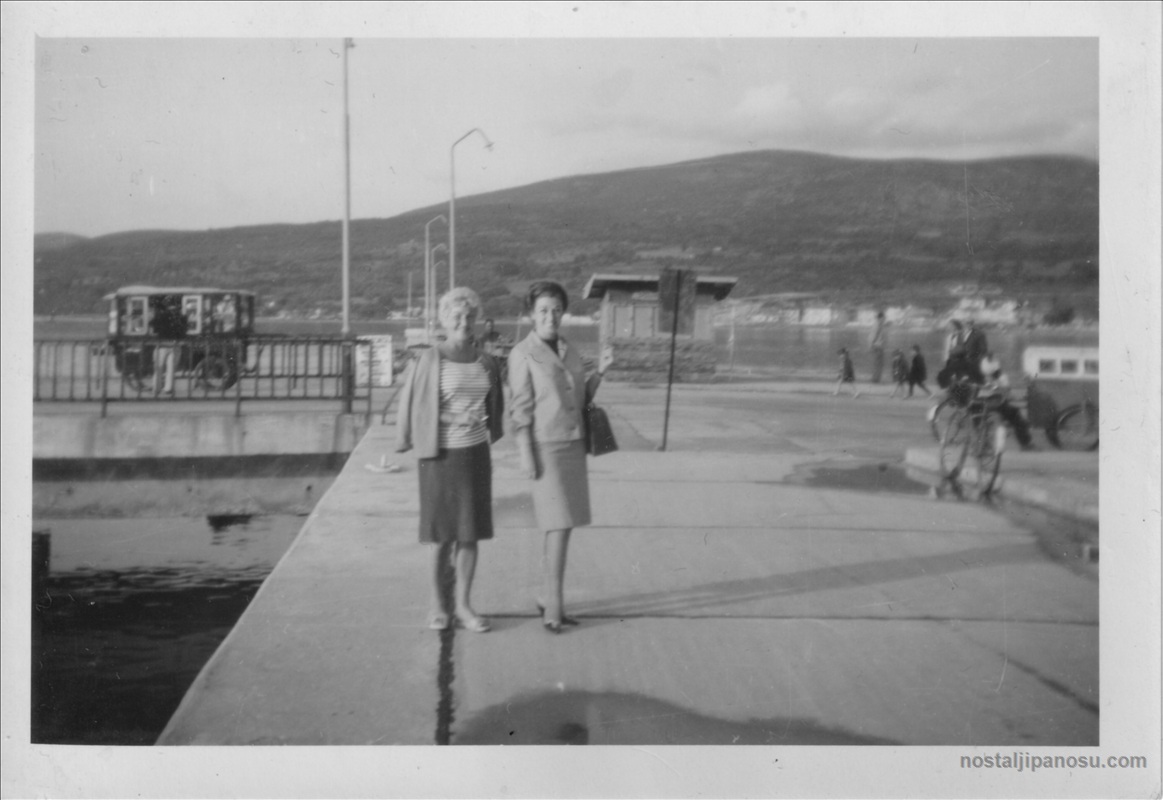1960'lı yıllarda iskelede duran, dönemin zarif döpiyes ve çizgili bluz modasını yansıtan iki kadının arkada dağ manzarasıyla siyah beyaz fotoğrafı.