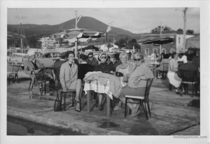 Sahildeki bir çay bahçesinde veya kafede, şemsiyelerin altında masalarda oturan gözlüklü ve şık kıyafetli insanların siyah beyaz karesi.