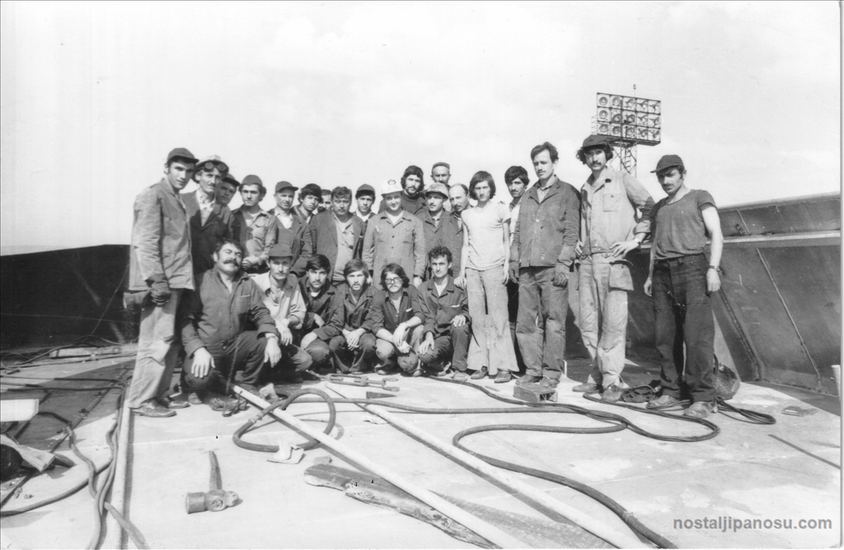 11 temmuz 1975 siyah beyaz tarihi işçi fotoğrafı; arka planda projektör kulesi, yerde inşaat kabloları ve aletleriyle poz veren bıyıklı, şapkalı tersane işçileri grubu.