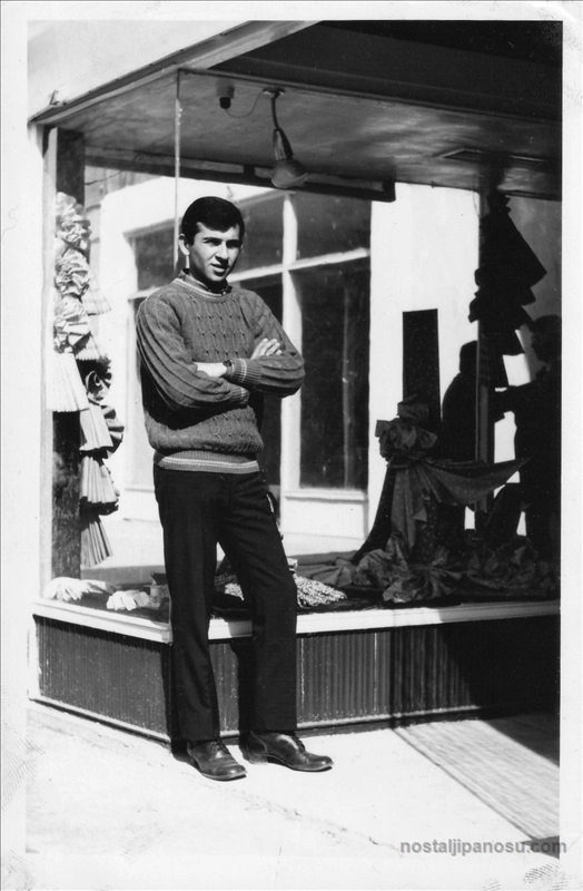 Nostaljik siyah beyaz fotoğrafta, retro örgü kazak giymiş genç bir adamın eski bir dükkan vitrini önünde kollarını kavuşturarak poz vermesi.