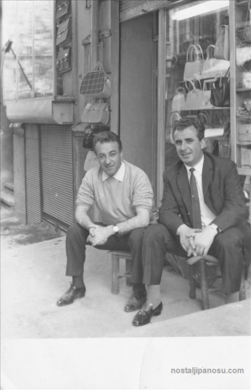 Siyah beyaz nostaljik fotoğrafta, eski bir çanta dükkanının önünde ahşap taburelerde oturan takım elbiseli ve kazaklı iki erkek esnaf.